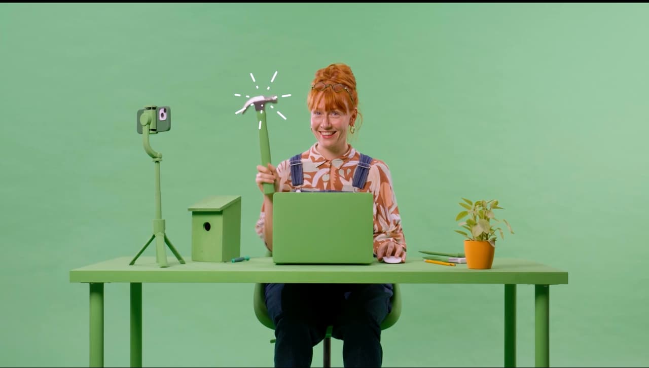 Red-haired woman holding a hammer at a green desk in an art-directed Awin studio set