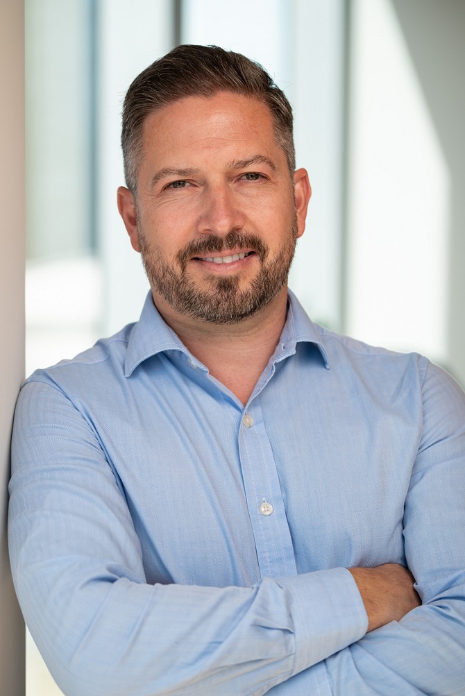 Male headshot smiling in front of large office window