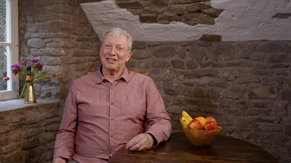 grey haired male sitting at table with fruit bowl smiling to camera