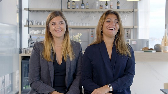 2 females sitting together in front of a bar smiling to camera