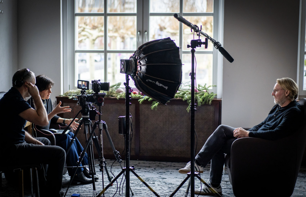 Corporate video interview being filmed with a professional camera, lighting, and microphone setup, showing a business leader in conversation with a video production team.