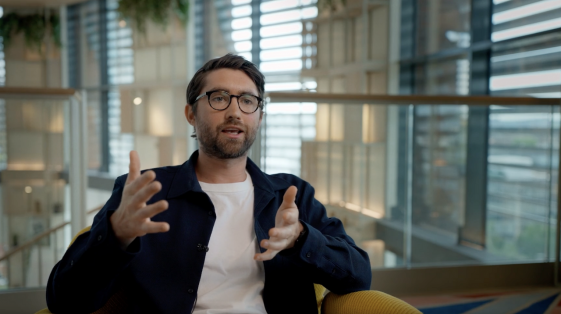 A confident man seated in a modern office, gesturing with his hands as he speaks directly into the camera, delivering a message with authority and focus."