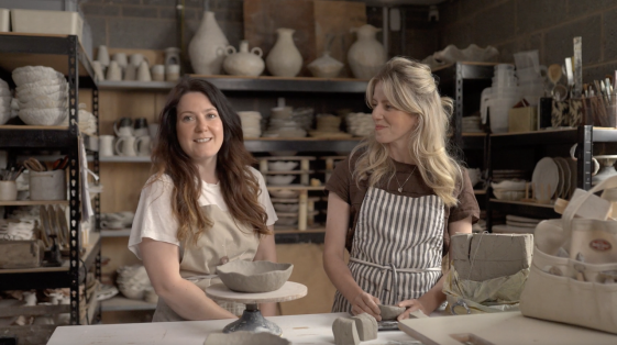 Two women smiling, with one facing the camera and the other looking at her friend who is speaking