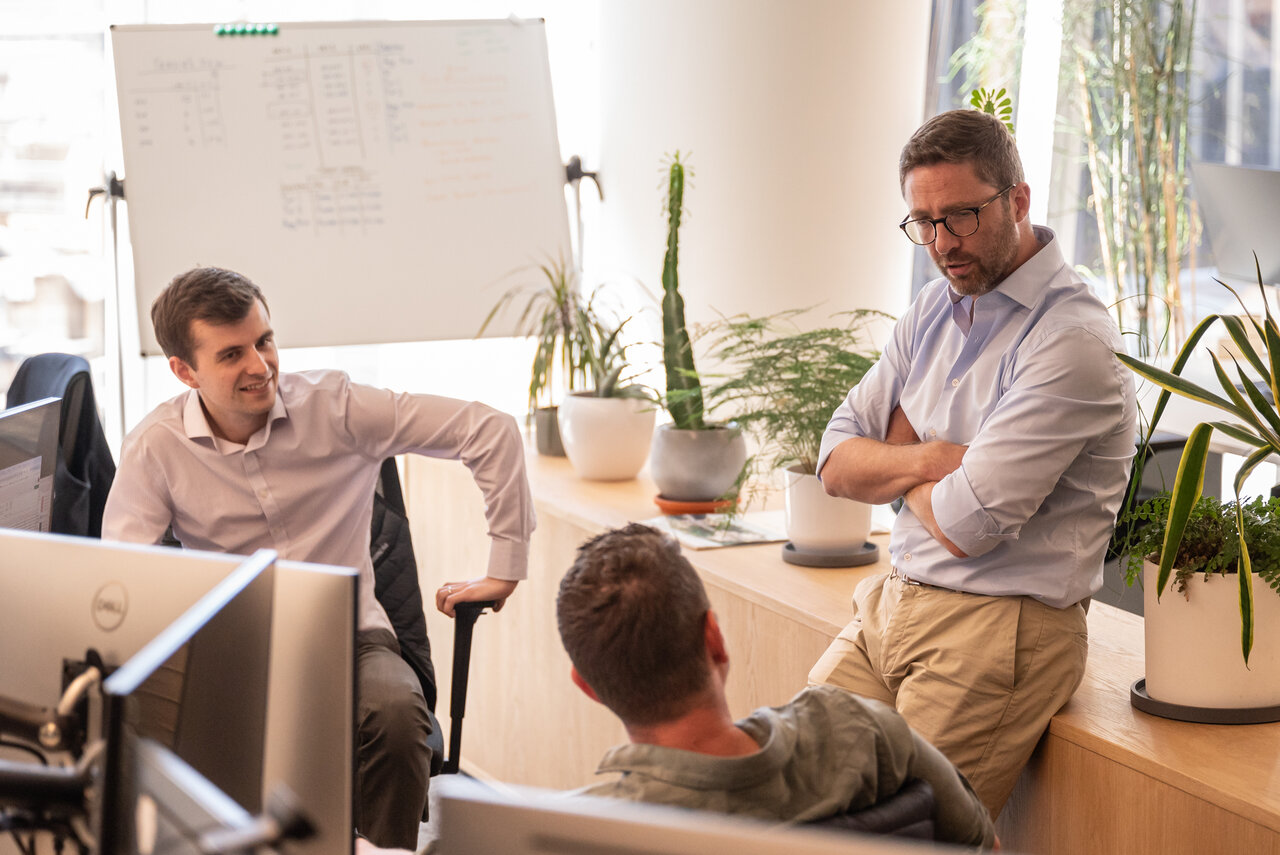 Three men in an informal meeting at desks in a corporate office setting.