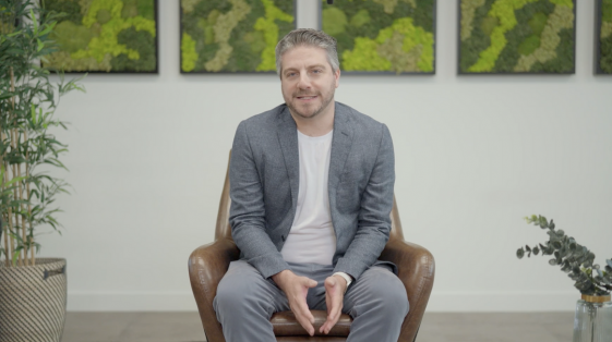 Man with grey hair sitting on a chair in the centre frame of a camera