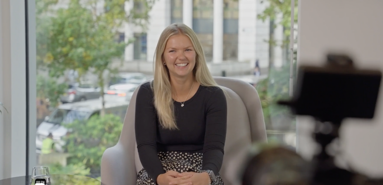 Smiling woman with long blonde hair in a black top and patterned skirt, seated by a window with trees.