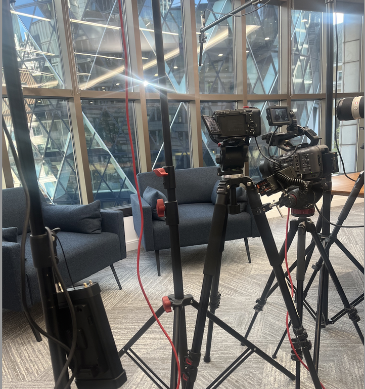 Video production setup with two cameras on tripods facing grey sofas in a glass-walled, sunlit studio.