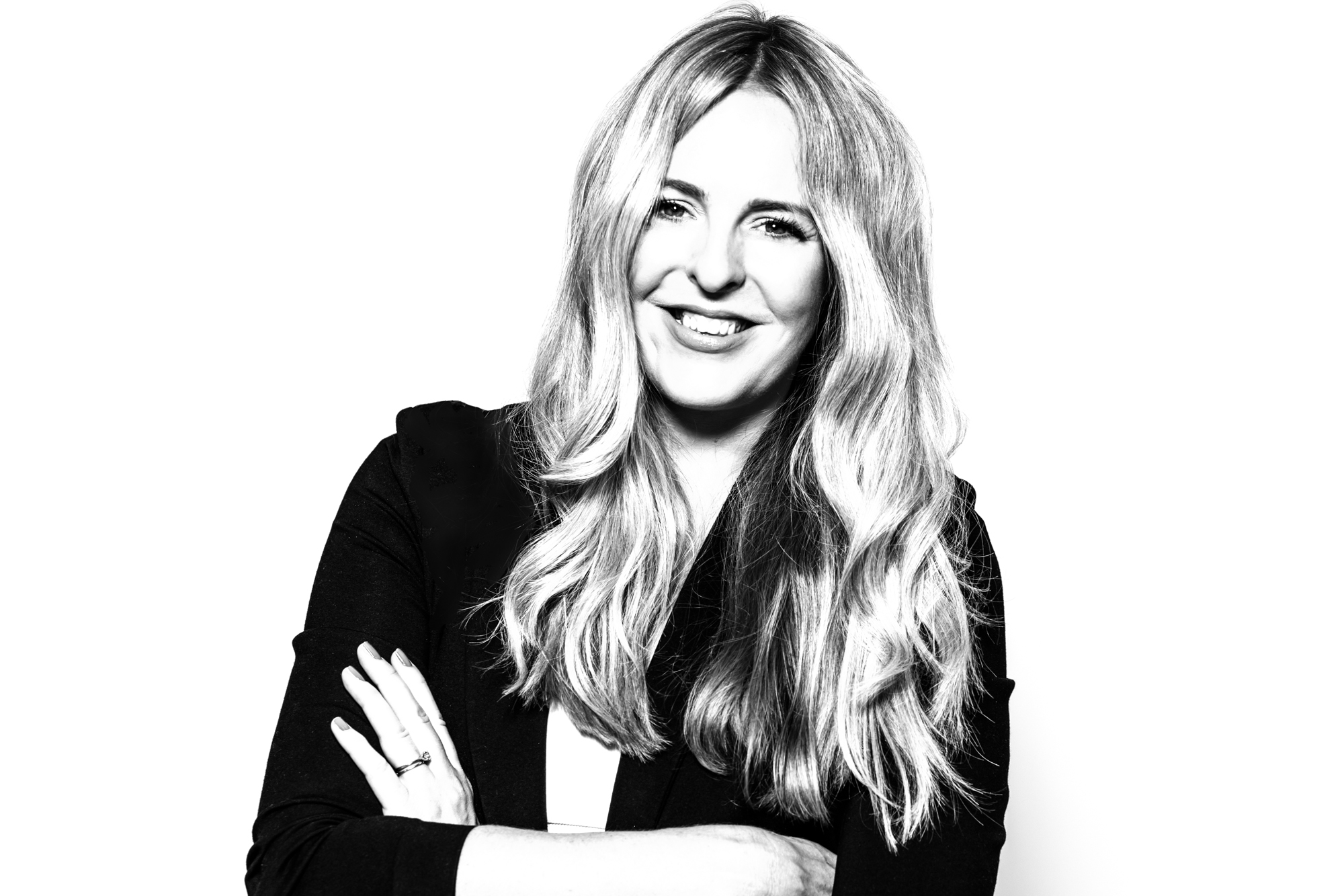 Black and white photo of a person with long, wavy hair smiling and standing with arms crossed. They're wearing a dark blazer over a light top, exuding the confident aura of a video production agency founder. The background is plain white.