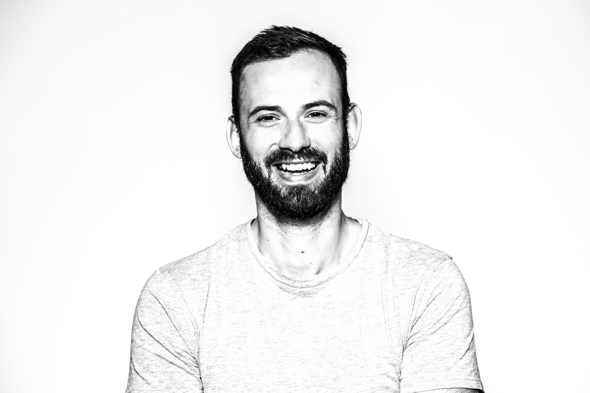 Black-and-white photo of a smiling man with a beard and short hair, wearing a light-colored t-shirt. His confident gaze reflects his role as the founder of a successful video production agency, set against a plain white background.