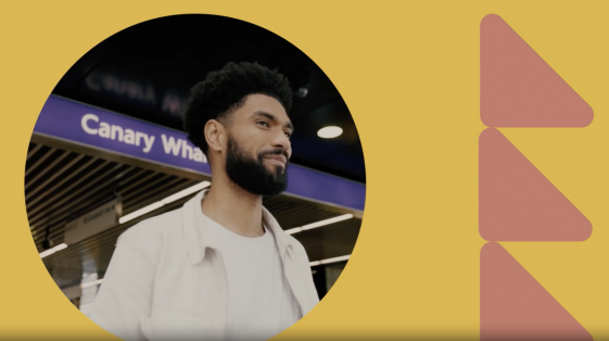 A man with a beard stands in front of a Canary Wharf London sign, smiling. The background is a vibrant yellow graphic enhanced by pink geometric shapes on the right.