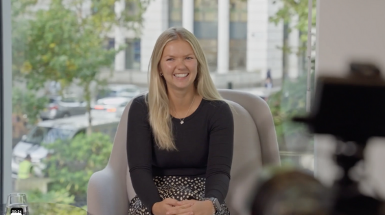 A woman with long blonde hair sits smiling in a comfortable chair, wearing a black top and patterned skirt. Behind her is a window with a view of trees and buildings. The scene, captured by a recruitment video production company, highlights the essence of their employer brand.
