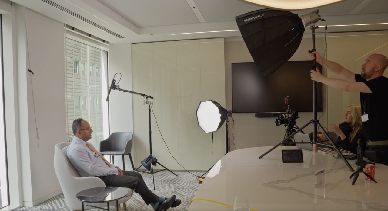 A man sits in a chair during a filmed interview, crafted by a recruitment video production agency. Bright studio lights and camera equipment surround him as two crew members adjust the setup. The setting exudes the ambiance of a modern workspace, perfectly tailored for impactful storytelling.