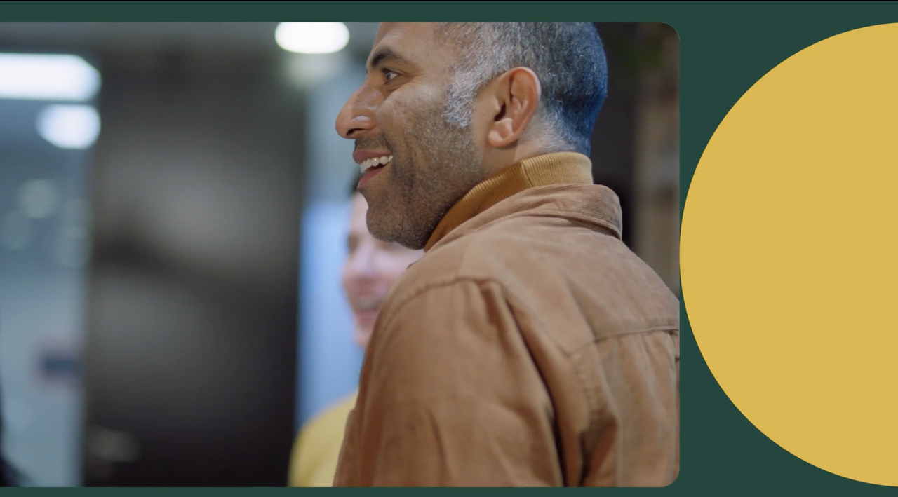 A man in a brown jacket smiles while standing indoors. The background is softly blurred, and a yellow circle is partially visible on the right side of the image.