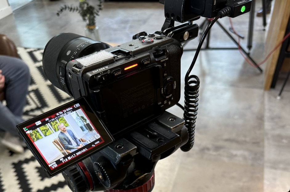 A digital camera, expertly positioned on a tripod by the video production agency, captures a video interview. The camera's screen displays a person seated on a chair, while equipment, cables, and part of a geometric-patterned rug contribute to the professional setup in the background.