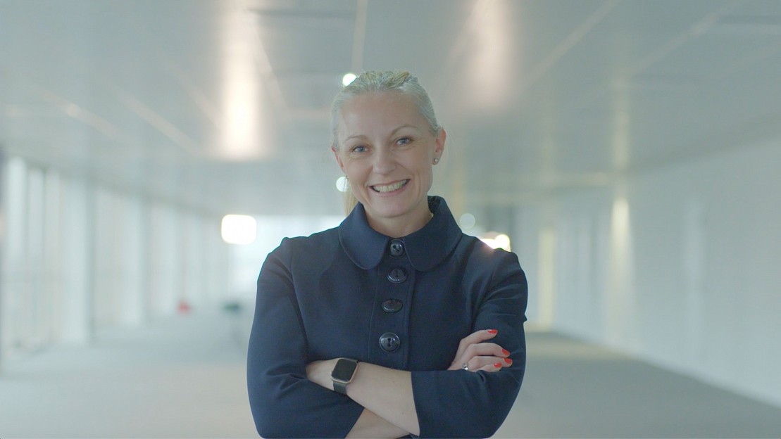 Female with blonde hair wearing a smart suit with arms crossed smiling to camera in the centre of a boardroom