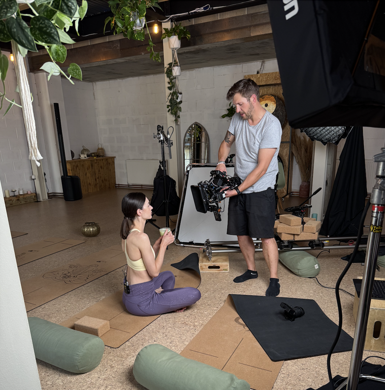 Behind-the-scenes photo of a video shoot in a yoga studio. A female actor in activewear sits on a yoga mat holding a drink, while a camera operator shows her the camera screen. Filming equipment and props like mats, lights, and yoga blocks are visible.