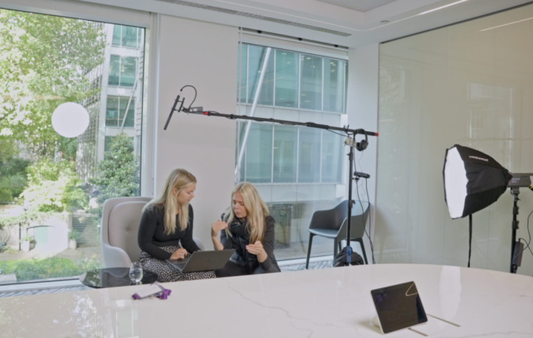 Behind the scenes of a corporate video shoot, showing two professionals discussing content while a video interview setup with lighting and audio equipment is prepared